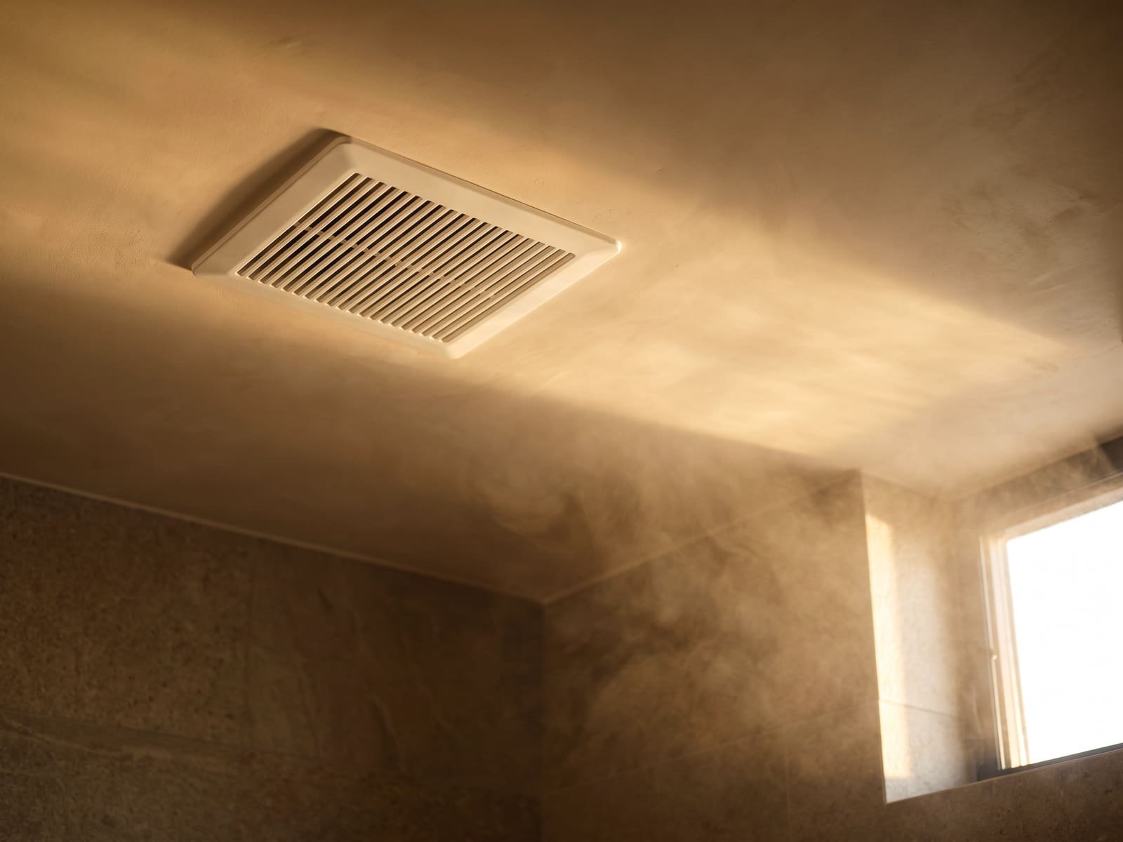 White ceiling exhaust fan grille pulling steam in a warm bathroom